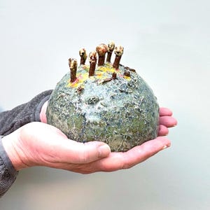 Könnte beinhalten: Eine handgefertigte Keramikskulptur mit einer strukturierten, kugelförmigen Form. Die Skulptur ist überwiegend grün mit gelben und roten Bereichen und weist mehrere braune, stielartige Vorsprünge auf der Oberseite auf. Das Stück wird in offenen Händen gehalten.