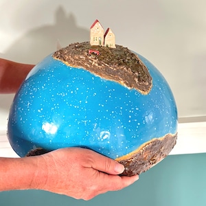 Può includere: Una scultura in ceramica a forma di cupola con una superficie blu brillante lucida e macchie bianche. Sulla parte superiore si trova una massa terrestre marrone testurizzata con due piccole case beige dai tetti rossi e una panchina rossa.