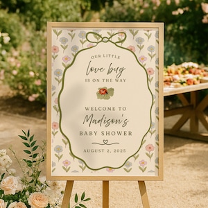 May include: A framed baby shower sign on an easel. The sign has a floral background with the text "Our Little Love Bug Is On The Way" and "Welcome to Madison's Baby Shower" with the date August 2, 2025.