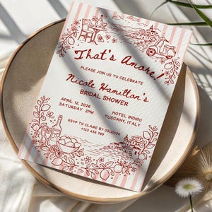 May include: A bridal shower invitation with the words "That's Amore" in a decorative font. The invitation features a red and white striped background and illustrations of Italian scenery. The invitation is on a beige plate.