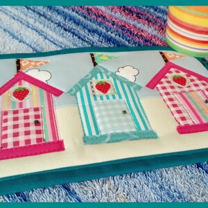 Op de afbeelding: Een stoffen placemat met drie kleurrijke strandhuisjes met aardbeienaccenten. De huisjes hebben roze, turquoise en witte strepen of ruitpatronen. De placemat is afgezet met teal en heeft een lichtblauwe hemel als achtergrond.