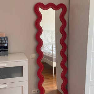 May include: A tall, rectangular mirror with a wavy, bright red frame. The mirror is positioned against a light-colored wall, reflecting a bedroom interior. The frame's unique design adds a decorative element to the room.