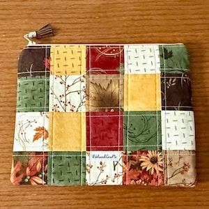 Puede incluir: Un estuche rectangular acolchado con un diseño de patchwork. Los cuadrados de tela presentan patrones con temas otoñales en marrón, verde, amarillo y rojo. Una pequeña borla cuelga de la cremallera.