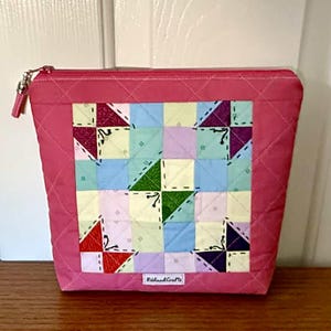Puede incluir: Un neceser rosa acolchado con un diseño de patchwork. El patchwork presenta cuadrados de varios colores, incluyendo azul, verde, morado y blanco, con un patrón de costura decorativo. La bolsa tiene un cierre de cremallera y una pequeña etiqueta que dice "Riland Crafts".