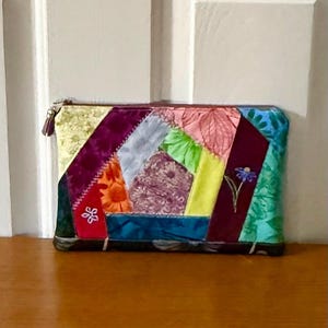 Puede incluir: Un bolso de mano patchwork con cierre de cremallera y borla. La bolsa rectangular presenta una variedad de parches de tela coloridos, incluidos patrones florales y geométricos. Se puede ver una pequeña flor bordada.