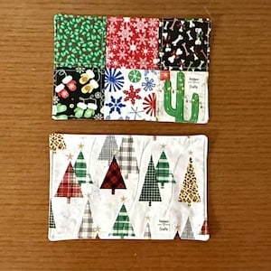 May include: Two rectangular fabric coasters. The top coaster has six patterned squares with Christmas and winter themes. The bottom coaster features Christmas trees in various patterns. The coasters are on a brown wooden surface.