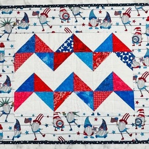 May include: A white quilt with a red, white and blue patchwork design. The quilt is made up of triangles and features a zig-zag pattern. The quilt is bordered with a black and white trim. The quilt is decorated with a variety of gnomes wearing red, white and blue clothing.