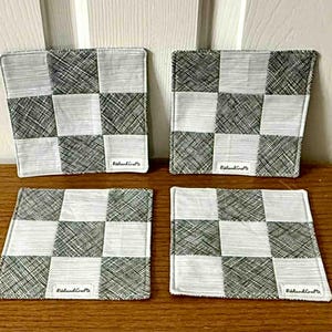 May include: Four square coasters with a black and white checkerboard pattern. Each coaster has a white label with the text "RolandCrafts". The coasters are made of fabric and are placed on a wooden surface.