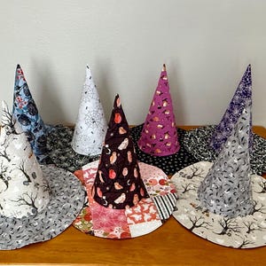 May include: A collection of eight fabric witch hats featuring various patterns including black trees, white spiderwebs, purple cauldrons, owls, and bats. The hats are conical with wide circular brims, showcasing a variety of colors like grey, white, black, purple, and orange.
