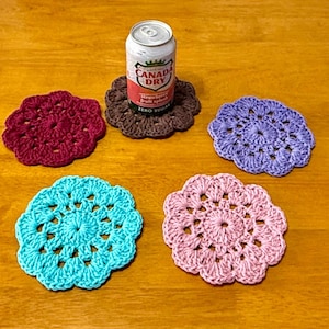 May include: Five round, crocheted coasters in maroon, brown, light purple, turquoise, and light pink. A silver and red Canada Dry Strawberry Fruit Splash soda can sits on the brown coaster. Each coaster features a scalloped edge and openwork.
