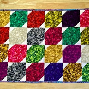 May include: A colourful quilt with a diamond pattern. The quilt is made of various fabrics with different patterns and colours, including red, green, purple, gold, and grey.