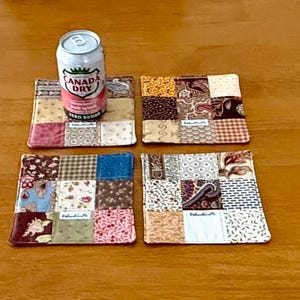 May include: A can of Canada Dry Zero Sugar soda sits atop a patchwork coaster. Four square coasters with various fabric patterns and colors are visible. The coasters are made of fabric squares in shades of pink, blue, brown, and cream.