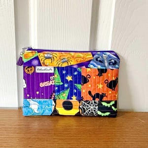 Peut inclure: Une pochette zippée sur le thème d'Halloween avec une variété de carrés de tissu colorés. Le motif comprend des araignées, des chats, des chauves-souris et le texte "IT'S GREAT". La pochette a une fermeture éclair violette et une pompon.
