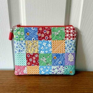 May include: A colorful patchwork zippered pouch with a red zipper and tassel. The pouch is made of small fabric squares in various patterns and colors, including floral, gingham, and geometric designs. The pouch is sitting on a wooden surface.
