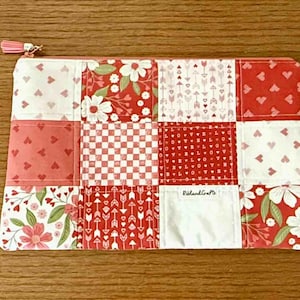 Puede incluir: Bolsa rectangular de patchwork con cierre de cremallera. El diseño presenta cuadrados rojos y blancos con patrones de corazones, flores, flechas y un diseño de tablero de ajedrez. El tirador de la cremallera es una borla rosa.