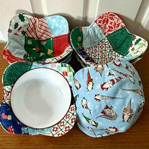 May include: A collection of four handmade bowl cozies in various Christmas-themed fabrics. The cozies feature patchwork designs with red, green, and blue patterns, including stars, gifts, and gnomes. One cozy holds a white bowl.