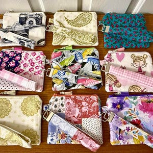 以下が含まれることがあります： 手作りのファスナー付きポーチのコレクション。リストストラップ付き。 カメラ、ペイズリー、花柄、漫画のスニーカーなど、さまざまなパターンが特徴です。 各ポーチには、お揃いのリストストラップと金属製のジッパーが付いています。