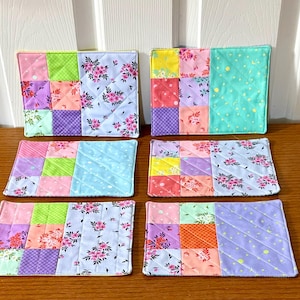 May include: Set of six colorful patchwork coasters. Each coaster features a unique combination of floral and geometric patterns in shades of pink, purple, green, and blue. The coasters are quilted and have a soft, padded appearance.