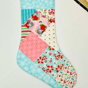 May include: A patchwork Christmas stocking with a blue, pink, and white color scheme. The stocking features a variety of holiday-themed patterns, including snowflakes, flowers, and cats.