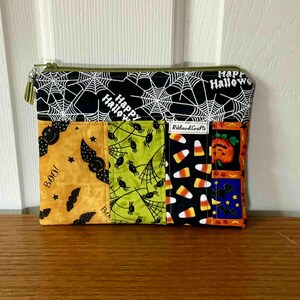May include: A rectangular Halloween-themed zipped pouch. The top section features a black and white spiderweb pattern with "Happy Halloween" text. The lower section has various Halloween-themed panels with bats, candy corn, and pumpkins. The zip is green.