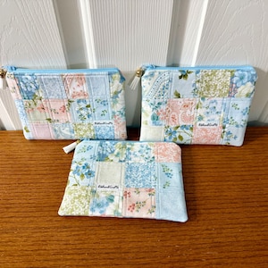 Puede incluir: Tres neceseres con cremallera de patchwork de diferentes tamaños. Presentan un fondo azul claro con estampados florales y geométricos en rosa, verde y blanco. Cada neceser tiene una cremallera azul claro y un tirador de borla. La marca "Ribbon&Crafts" es visible.