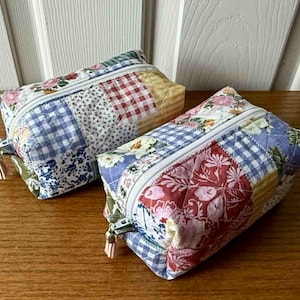May include: Two rectangular quilted zipper pouches. One features a patchwork design with floral, gingham, and solid fabric squares in blue, white, yellow, and red. The other has a similar design with red floral and gingham patterns.
