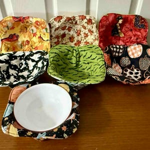 May include: A collection of fabric bowl cozies in various Halloween-themed patterns. Designs include ghosts, bats, pumpkins, and witch illustrations. One cozy holds a white ceramic bowl. The cozies are displayed on a wooden surface.