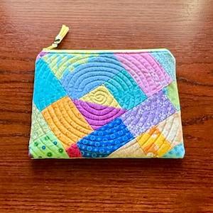 May include: A colorful, quilted pouch with a zipper closure. The patchwork design features various geometric shapes in shades of blue, green, yellow, purple, and orange. The zipper pull is yellow.