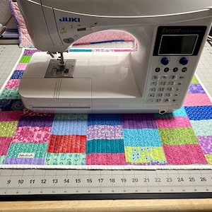 May include: A white Juki Exceed sewing machine rests on a patchwork quilt with squares of pink, blue, green, and purple fabrics. The machine has a digital display and control buttons. The quilt is on a gray cutting mat with measurements in inches.