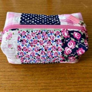 May include: A small, quilted cosmetic bag with a pink zipper and tassel. The bag features a patchwork design with floral, polka dot, and gingham patterns in pink, white, and navy blue. The bag is approximately 6 inches long.