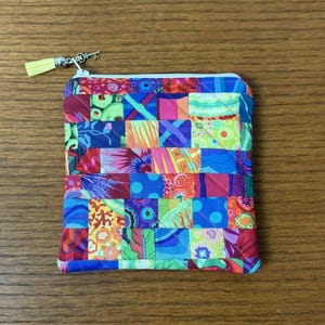Könnte beinhalten: Ein kleiner, quadratischer, gesteppter Beutel mit einem farbenfrohen Patchwork-Design. Der Beutel hat einen Reißverschluss und eine gelbe Quaste am Reißverschlussanhänger. Das Patchwork enthält verschiedene Farben und Muster, darunter Blumen- und geometrische Designs.