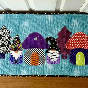 Könnte beinhalten: Halloween-Tischläufer mit applizierten Zwergenhäusern in Pilz- und Baumform. Die Häuser haben verschiedene Muster, darunter Punkte, Spinnweben und Schachbrettmuster. Der Hintergrund ist ein hellblau gesteppter Stoff.