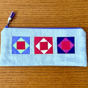 Peut inclure: Une trousse rectangulaire bleu clair avec une fermeture éclair et un pompon violets. La trousse présente trois blocs de courtepointe carrés rouges, blancs, verts et violets. La trousse mesure environ 23 cm de long.