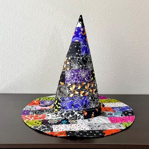 May include: A conical hat featuring a patchwork design of various Halloween-themed fabrics. The patterns include spider webs, candy corn, ghosts, and moons in colors of purple, orange, black, white, and green. The hat has a wide, flat circular brim.
