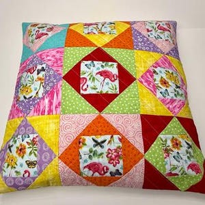 May include: A colorful patchwork pillow with a square design featuring pink flamingos, butterflies, and floral patterns. The pillow is made of various fabrics in shades of pink, yellow, green, red, and purple.