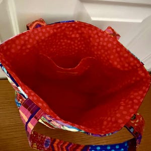 以下が含まれることがあります： 鮮やかな赤色の内装と小さなポケットが付いた、カラフルなトートバッグ。バッグの外側は、青、オレンジ、紫など、さまざまな模様と色を使ったパッチワークデザインです。ハンドルも模様入りです。