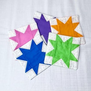 May include: Five fabric coasters featuring a star design. The coasters are white with pink, purple, orange, green, and blue stars in the centre. The stars are surrounded by a white border with small pink dots.