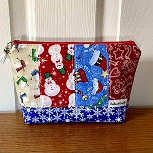 Puede incluir: Un neceser navideño de patchwork con cremallera. Presenta paneles con muñecos de nieve, gorros de Papá Noel y hombrecitos de jengibre. La base es azul con copos de nieve blancos. Cremallera roja.