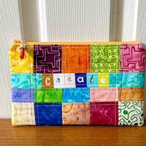 May include: A colorful patchwork zippered pouch with the word "CREATE" stitched across the front. The pouch features a variety of patterned fabric squares in shades of yellow, purple, orange, green, and pink. A yellow zipper and tassel complete the design.