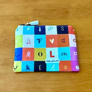 May include: A colorful zippered pouch with a patchwork design featuring letters of the alphabet. The pouch has a light blue and white tassel on the zipper pull. The letters are in various colors and sizes.
