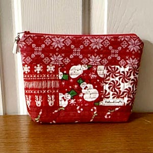 Puede incluir: Bolsa roja con cremallera con temática navideña y varios diseños. Los diseños incluyen copos de nieve, muñecos de nieve, bastones de caramelo y un diseño de punto. La bolsa es de tela.