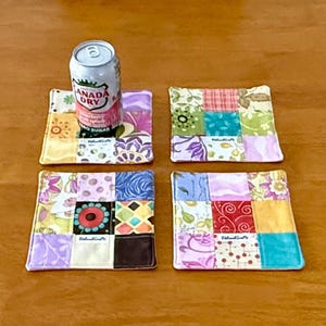 May include: Four square fabric coasters with a patchwork design in various colors and patterns. A can of Canada Dry ginger ale sits on one coaster. The coasters are on a wooden surface.