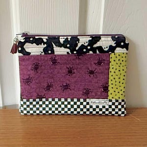May include: A rectangular, handmade zippered pouch with a patchwork design. The top section features a black and white pattern, the middle a purple fabric with insect motifs, and the side a green dotted fabric. The bottom has a black and white checkered pattern.