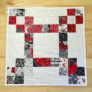 May include: A white quilted table runner featuring a red and black Christmas design. The runner is made from fabric squares with various patterns, including holly, baubles, and snowflakes.