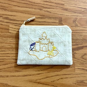 Peut inclure: Pochette matelassée de couleur crème avec une fermeture éclair et un pompon dorés. La pochette présente une scène de château de sable brodée avec un seau bleu et une pelle jaune. Le motif est cousu en fil d'or et bleu.
