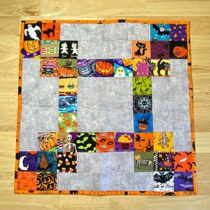 May include: A Halloween-themed patchwork quilt with a grey background and orange, black, and white fabric squares featuring various Halloween motifs, including pumpkins, cats, ghosts, bats, and candy corn. The quilt is edged with an orange border.