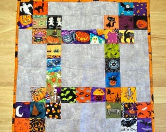 Pode incluir: Uma colcha de retalhos com tema de Halloween com fundo cinza e quadrados de tecido laranja, preto e branco com vários motivos de Halloween, incluindo abóboras, gatos, fantasmas, morcegos e milho doce. A colcha é bordada com uma borda laranja.