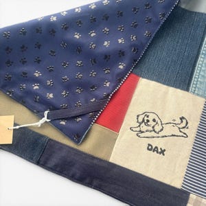 May include: A patchwork pet bandana featuring a navy blue fabric with a repeating black paw print pattern, combined with panels of denim, red, and tan fabric. A tan square panel displays a black line drawing of a dog with the name DAX below it.