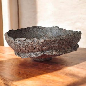 Puede incluir: Un cuenco texturizado y hecho a mano con un aspecto rugoso y natural. El cuenco es de color marrón oscuro y terroso y se asienta sobre una pequeña base redondeada. El borde tiene un borde irregular y dentado, lo que realza su estética única. Este cuenco decorativo es adecuado para la decoración del hogar.