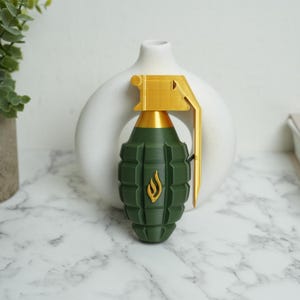 May include: A green and gold decorative grenade with a flame emblem. The grenade features a textured body and a gold-colored lever and top. The background includes a white vase and a marble surface.